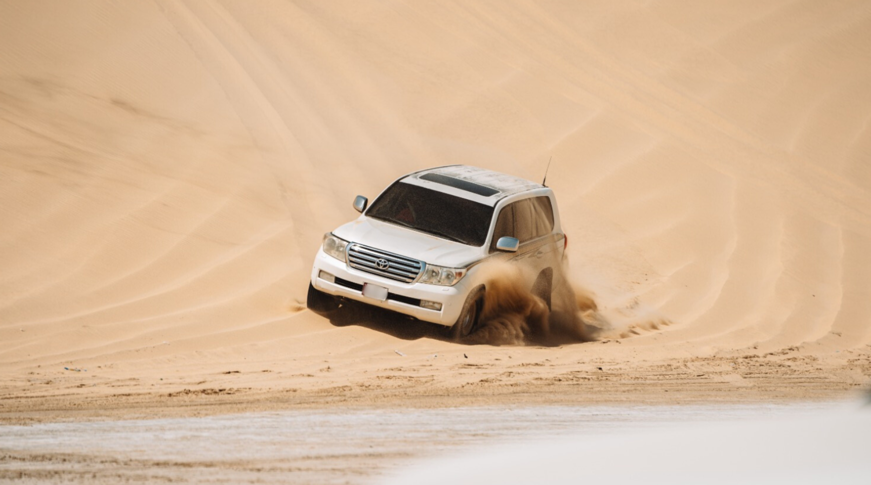 A white SUV navigating through sandy dunes under a clear blue sky.