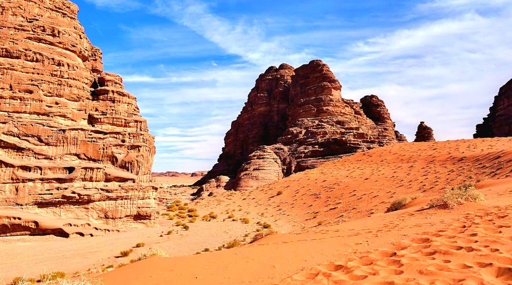 A vast desert landscape featuring red sand and striking rock formations under a clear blue sky.