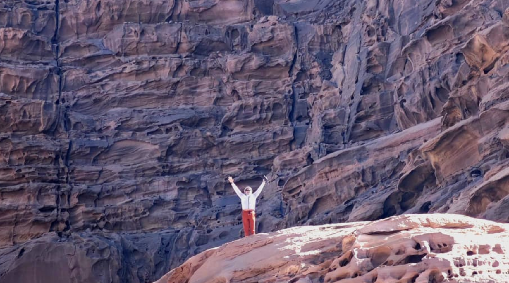 A person stands triumphantly on a mountain peak, arms raised in celebration against a clear blue sky.