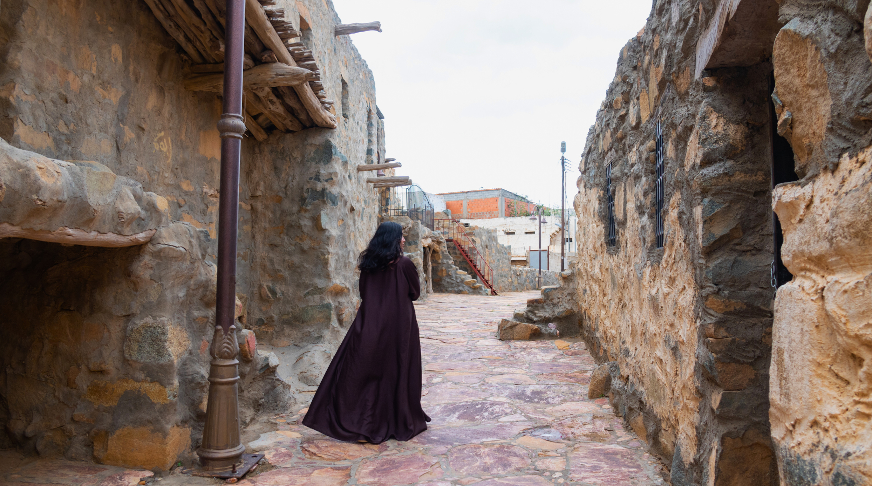 A woman in a flowing dress strolls through a narrow alley, surrounded by brick walls and soft shadows.