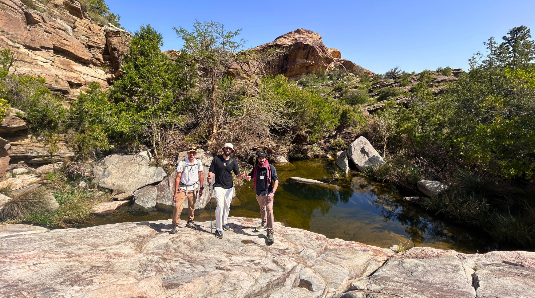 Three people stand on rocks beside a flowing stream, surrounded by greenery and natural scenery.